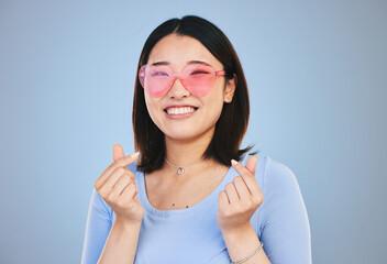 Happy asian woman, portrait and sunglasses with love hands, sign or gesture against a blue studio...
