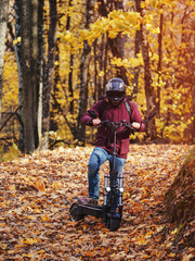 Fototapeta premium A young man rides a scooter in the autumn park. Powerful modern electric transport. Autumn road in maple leaves
