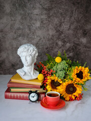 plaster copy of classic ancient bust, flowers, tea cup, alarm clock, stack of books close up on table, abstract dark background. back to school concept. autumn time. Knowledge Day, education
