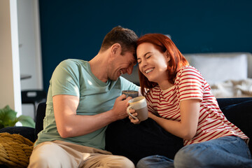 Happy carefree middle aged couple in love joking laughing while relaxing on couch with coffee, bonding, enjoying morning time together at home. Emotional connection in a relationship. Gladness concept