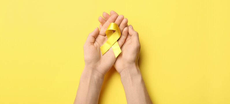 Hands with golden awareness ribbon on yellow background. Cancer concept