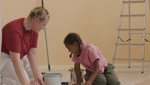 Multiracial mother and daughter sitting on floor covered with polythene sheeting in renovating apartment and mixing paint in plastic can