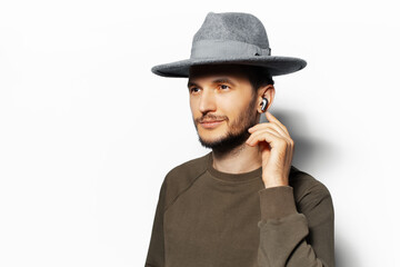 Studio portrait of young man touching the wireless earphone in his ears, wearing green sweater and grey hat isolated on white background.