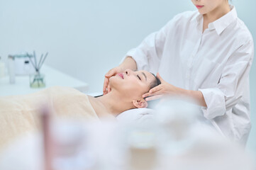 아시아 한국의 젊은 여성이 스파 또는 피부관리전문점 또는 피부과 병원에서 침대에 누워 베이지색 대형 타올을 덮고 눈을 감고있고 하얀색 셔츠를 입은 전문관리사가 손으로 얼굴 피부관리 또는 두피관리를 받고 있다. 