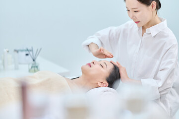 아시아 한국의 젊은 여성이 스파 또는 피부관리전문점 또는 피부과 병원에서 침대에 누워 베이지색 대형 타올을 덮고 눈을 감고있고 하얀색 셔츠를 입은 전문관리사가 손으로 얼굴 피부관리 또는 두피관리를 받고 있다. 