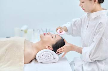 아시아 한국의 젊은 여성이 스파 또는 피부관리전문점 또는 피부과 병원의 침대에 누워 베이지색 대형 타올을 덮고 눈을 감고있고 하얀색 셔츠를 입은 전문관리사가 손에 분홍색 클렌징 스펀지를 들고 얼굴 피부관리를 하고 있다. 