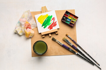 Mini easel with paints, brushes and glass of water on white grunge background