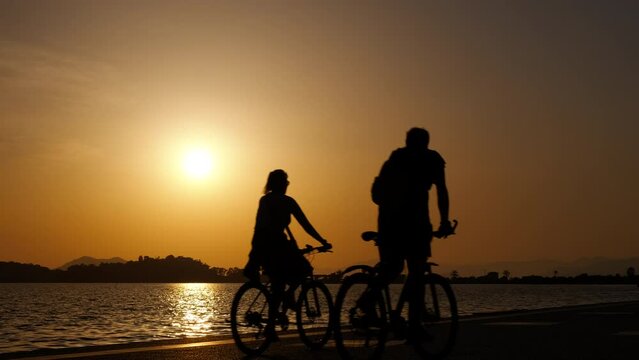 Silhouette of couple on bikes at lake sunset. A view of lovely couple silhouettes rides bikes in the city strees by the evening river in summer. A concept of active promenade on the river bank.