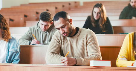 Young handsome African American male student listening lection at college, writing down and using textbook.