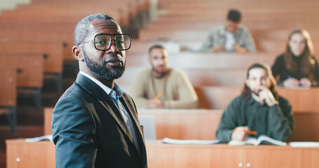 Rear of African American male professor standing in front of students in college and talking to...