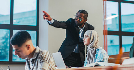 African American man professor walking the auditorium during exam and helping multiethnic students...