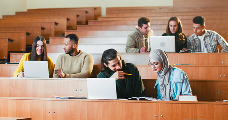Mixed-races males and females students sitting in auditorium with laptops, talking and working on task at college. Multiethnic guys and girls at computers discussing studying project at high school.