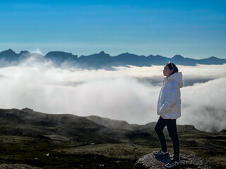 A young woman standing and enjoying the view of misty clouds covering the mountain range in front. in cold weather and has a beautiful view