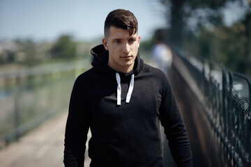 Young Man with black sweater walk outside in park in midday sun and look down to side with a neutral and serious face expression