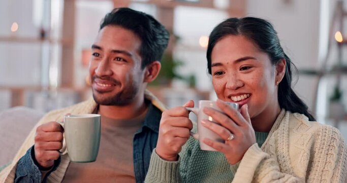 Coffee, happy and couple with blanket on sofa for bonding, relax and cozy in living room with fire. Love, dating and Asian man and woman with hot drink in conversation, talking and speaking at home