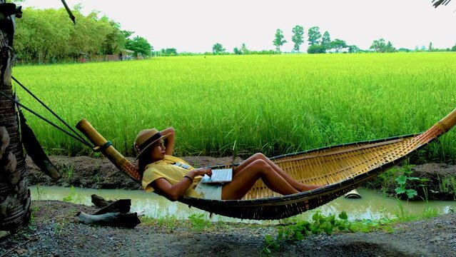 Asian women in a hammock working on a laptop with on the background green rice paddy fields in Thailand
