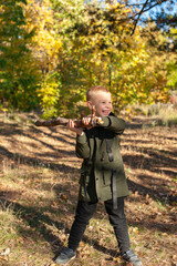 Naklejka premium A little boy in a green jacket plays with a stick in nature in the autumn forest