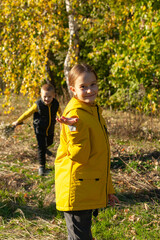 Brother and sister are walking in the autumn forest.  Girl looking at the camera. The girl is dressed in a yellow jacket, the boy is wearing a black vest. 