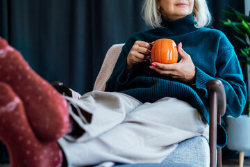 Cropped mature middle aged woman in sweater and warm socks relaxing in armchair with pumpkin shaped cup of hot coffee or tea drink. Cozy calm autumn holidays at home. Fall hygge mood concept.