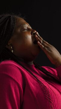 vertical video Female fashion influencer in isolated black background yawning to show her fatigue after hard day. Side-view portrait of fashionable african american woman covering her mouth.