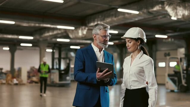 Two Engineers Using Tablet and Talking While Standing in the Factory Hall. Man and Woman Industrial Engineers in Hard Hats Discussing a New Project while Using a Tablet Computer