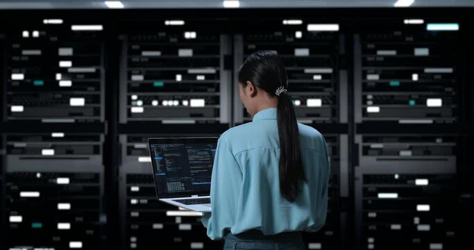 Back View Of A Woman Using A Laptop Writing Code In Server Room Database. Showing Database On Terminal Window In Dark Digital High Tech Room
