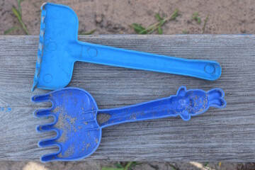 two small blue plastic toy rake lie on a wooden sandbox board outside in the playground