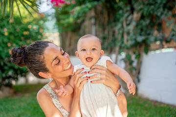 young mom holds her 6 month old baby in her arms while laughing with her mouth open in a garden with flowers and plants in the background. maternity and newborn baby care concept