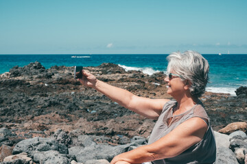 Happy senior woman with sunglasses enjoying sea vacation sitting at the beach in a sunny day taking a selfie with mobile phone