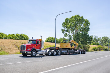 Red day cab powerful big rig semi truck tractor transporting excavator on step down semi trailer...