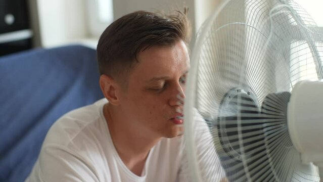 Tired man suffering from summer heat trying to cool off with eclectic fan at home. Sweaty male sit on sofa in front of ventilator cooling with electric fan cooler wind blowing without air conditioner
