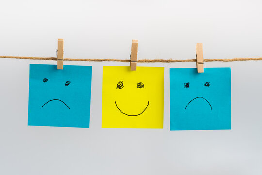 Positive attitude concept, three sticky notes hanging on a string attached with wooden clothespins, two sad faces in blue color and one standing out smiling face in yellow color in the middle.
