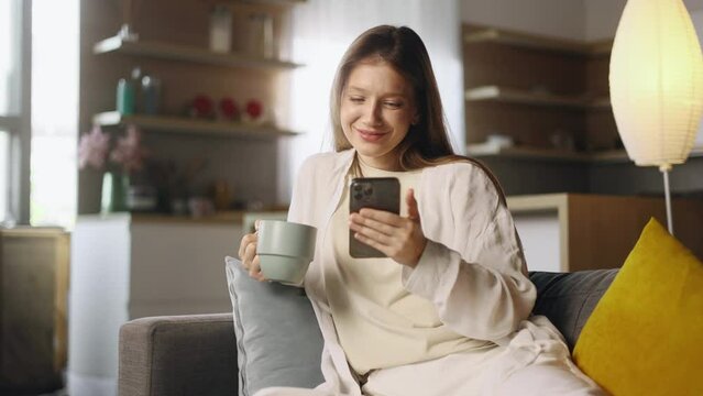 Charming young woman with cup of hot coffee or tea hold smartphone browsing scrolling app watching social media feed indoors Happy female texting on her phone enjoying leisure time at cozy home