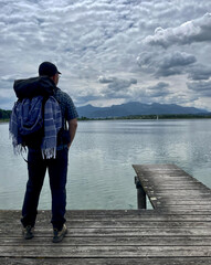 Mann mit Rucksack von hinten steht auf einem Steg am Chiemsee und schaut über den See in die...