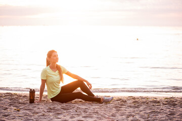 Fototapeta na wymiar Young woman in sportswear is sitting on the seashore at sunrise. Girl is resting after workout