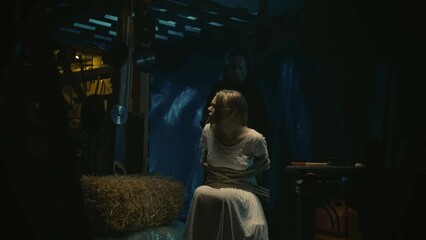 Fototapeta premium Girl victim tied with a rope sitting on a chair, man in the hockey mask with a chainsaw walking behind her.