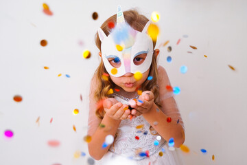 A child in a carnival mask of a unicorn, hand-made from glitter foamiran and faur fux. Little cute cheerful comical girl in the role of a unicorn, preparing for the carnival, masquerade, holidays.