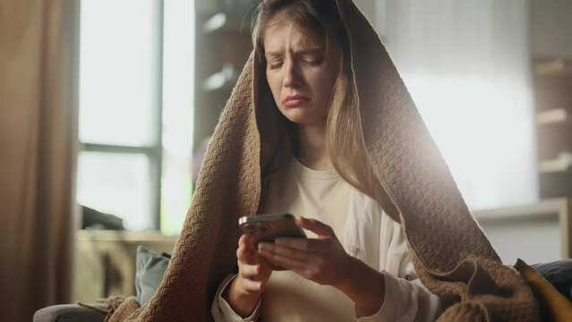 Portrait of crying young woman with smartphone sitting on the couch under the blanket and reading bad news email break up message from ex-boyfriend or personal problems at home