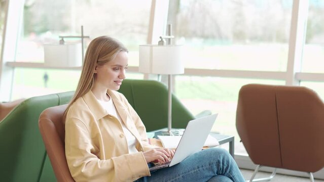Smiling young woman, teen girl student using laptop computer sitting on chair in university campus, coworking or airport lounge space hybrid learning or working online, browsing web, surfing.