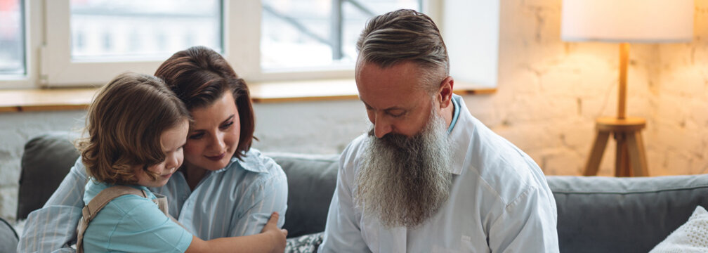 Doctor doing regular checkup in hospital. Sick little girl receive medical care. Senior male paediatrician examining sick kid sitting on mother's lap with stethoscope during home visit. Banner