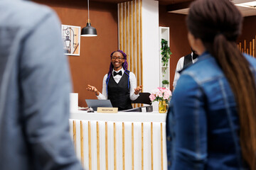 Friendly young African American female receptionist standing at hotel reception greeting tourists. Positive black girl front desk agent wearing uniform smiling to arriving guests
