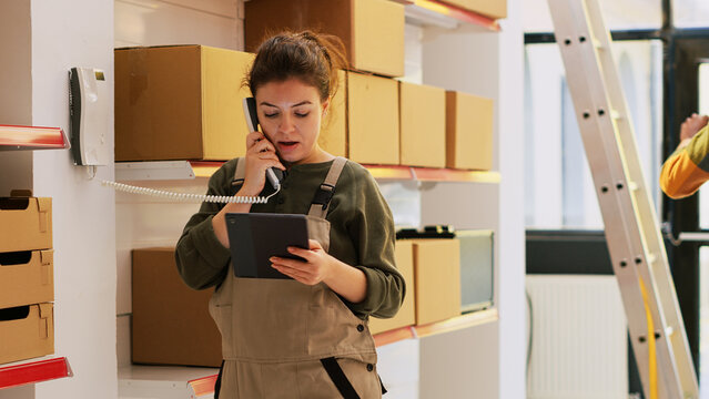 Manager talking to someone in storehouse, answering landline phone call to discuss with dispatcher about cargo distribution and production. Female supervisor inspecting merchandise.