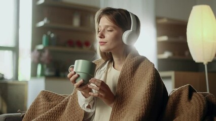 Portrait of pretty young woman under the blanket listening to music on sofa with soft light behind her indoors Smiling female with headphones hold cup of hot drink enjoying leisure time at home alone
