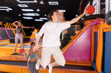 Dynamic young man throwing ball in basket while high jumping in trampoline arena