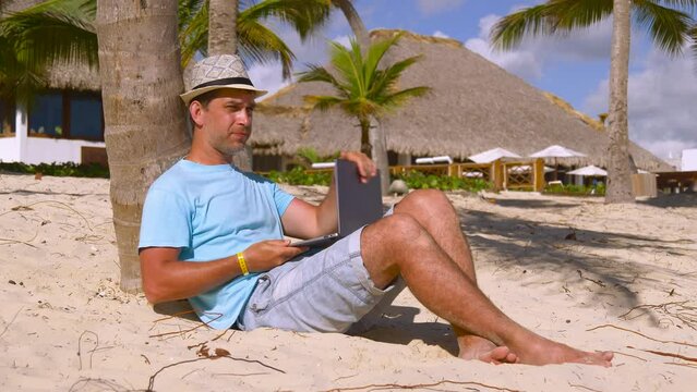 Freelancer on sand beach with notebook. Freelancer at work on beach under a palm tree. Freelancer finishes work, closes his laptop and exits the frame. Work and vacation. Freelance travel concept.