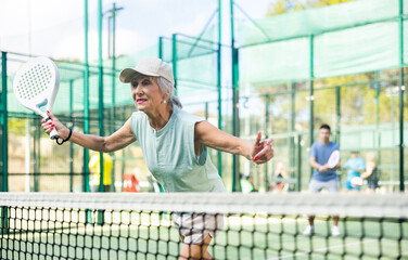 Sporty mature woman padel player hitting ball with a racket on a hard court