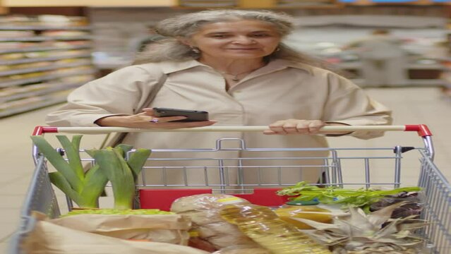 Vertical shot of senior woman working as mystery shopper walking along aisle in supermarket taking photos on smartphone, word on background translated as fish