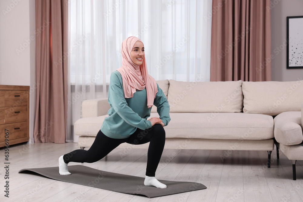 Wall mural Muslim woman in hijab doing exercise on fitness mat at home. Space for text