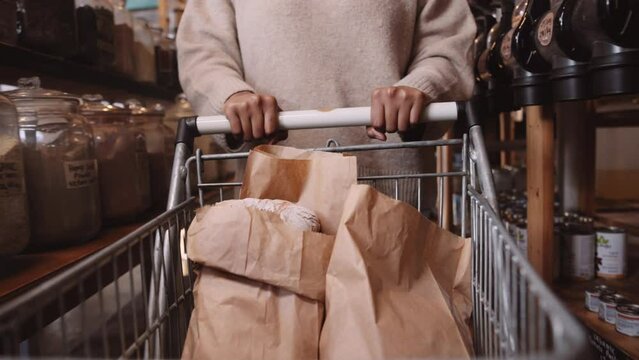 Close-up of young black woman pushing shopping trolley of paper bags in aisle of greengrocer's shop