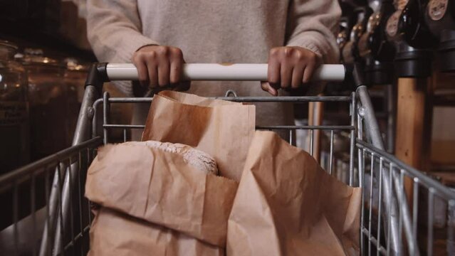 Close-up of young black woman pushing trolley of paper bags in aisle of greengrocer's shop